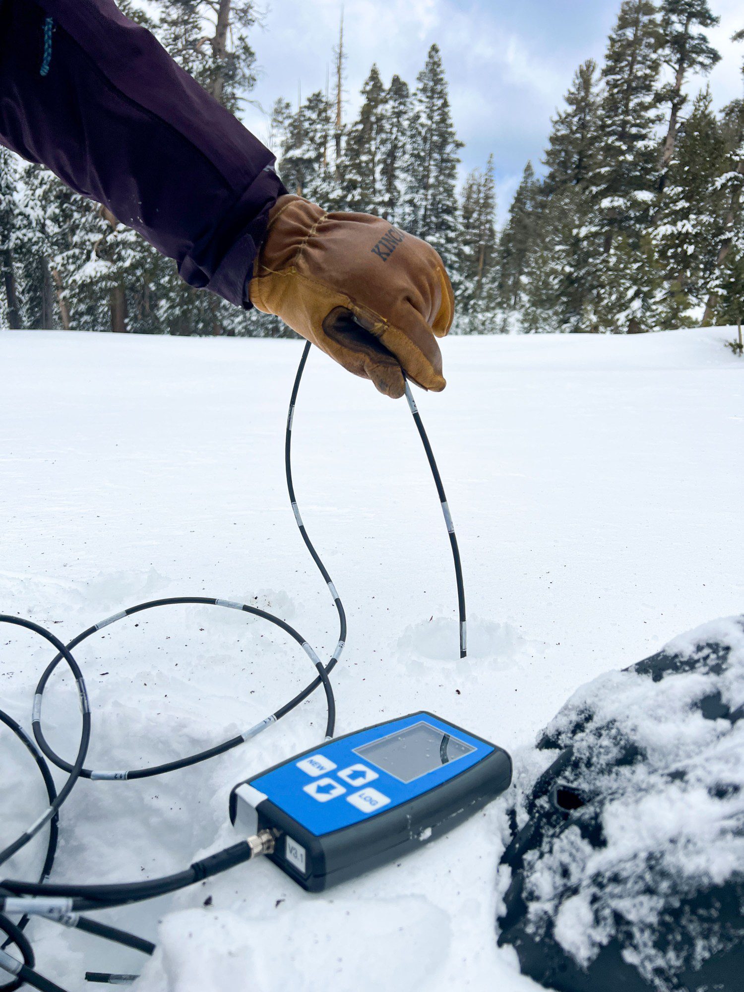a gloved hand inserts a probe wire into a hole in the snow