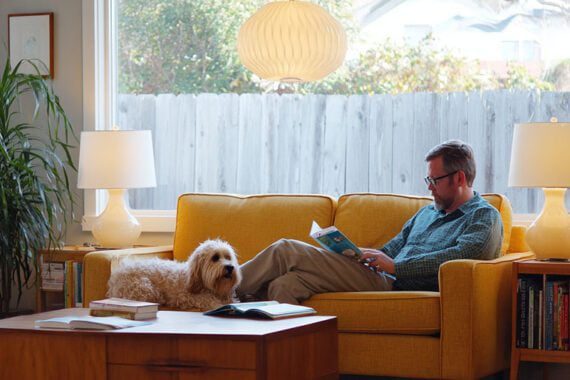 A man reading a book on a couch next to a dog