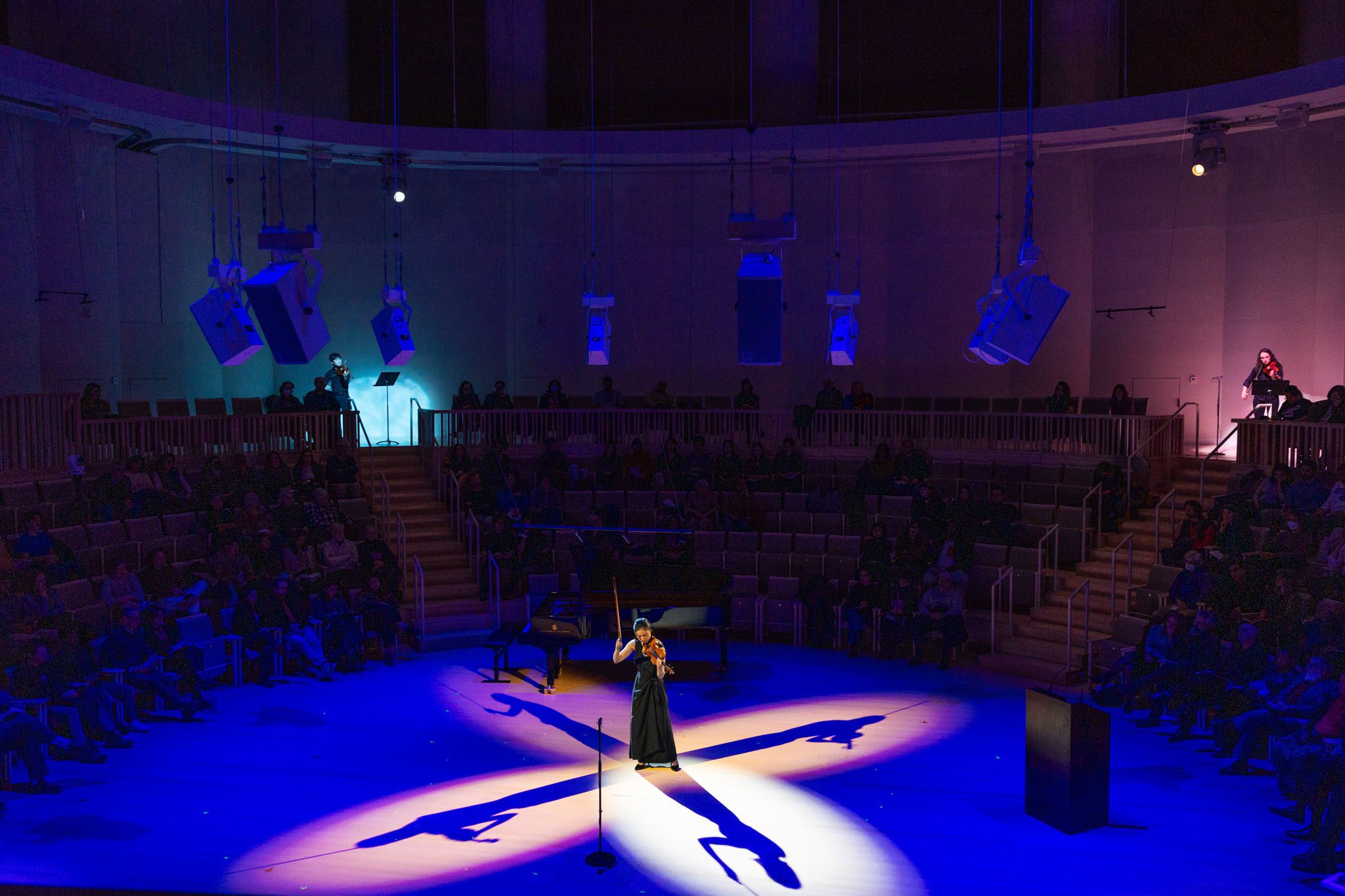 a woman holds her bow aloft as she plays the violin at the center of converging beams of the spotlights such that four shadows extend away from her at each 90 degree angle.